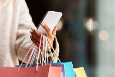 Tüketim ve Teknoloji. Tanımlanamayan siyah bir kadının akıllı telefon kullanması, alışveriş torbalarını tutması, dışarıda durması, kıyafet seçip sipariş etmesi, hediyeler alması, fotokopi alanı, bulanık arka plan.