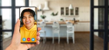 Bir kişi elinde bir akıllı telefon tutuyor ve bir video görüşmesinde gülümseyen bir bireyi gösteriyor. Arka planda modern bir mutfağı olan rahat bir ev..