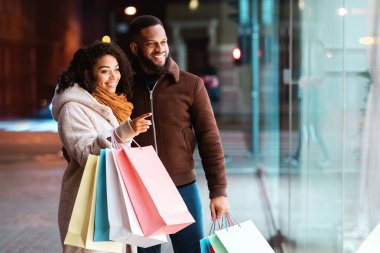 Şuna bir bak. Gülümseyen Afro-Amerikalı kadın erkeğine moda kıyafetlerini gösteriyor, vitrini işaret ediyor, akşamları alışveriş merkezinin yakınında yürüyor. Kış Satış ve İndirim, Perakende, Satın Alım