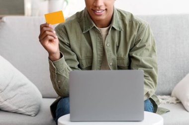 İnternetten alışveriş. Evden internet üzerinden mal almak için kredi kartı ve laptop kullanan neşeli siyahi bir gencin portresi. Covid-19 tecridi sırasında internetten bir şeyler alan Afro-Amerikalı genç adam.