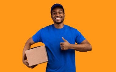 En İyi Teslimat Hizmeti Konsepti. Mavi şapkalı ve üniformalı gülümseyen siyah kurye elinde karton kutuyla sarı turuncu stüdyo arka planında izole bir şekilde duran baş parmak hareketlerini gösteriyor.