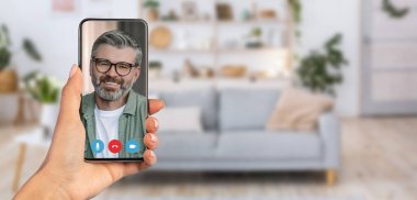 Bir kişi kamerada gülümseyen bir adamı gösteren bir akıllı telefon tutuyor. Arka plan, rahat bir oturma odası ortamı sunuyor. Kanepe ve dekorla. İletişim için rahat bir ortam..