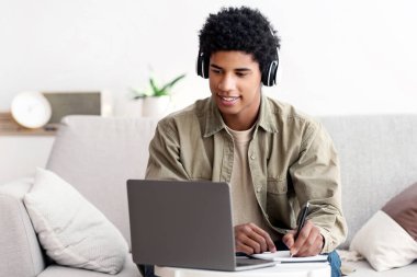 Web tabanlı eğitim konsepti. Kulaklıklı mutlu genç adam dizüstü bilgisayardaki öğretmenle çevrimiçi iletişim kurarken not alıyor. Çevrimiçi kursu için ödev yapan Afro-Amerikan ergen.