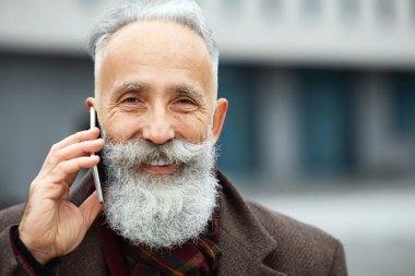 Sıcak kıyafetler içinde, sokakta yürürken cep telefonuyla konuşan neşeli, yaşlı bir adamın yakın plan portresi. Yakışıklı, sakallı, yaşlı iş adamı dışarıda akıllı telefondan konuşuyor.
