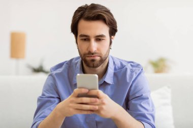 Ciddi Adam akıllı telefon kullanıyor kulaklık takıyor internete takıyor ve evdeki koltukta oturup internetten haber okuyor. Oturma odasında cep telefonundan mobil kullanıcı mesajı. Telefon Bağımlılığı Kavramı