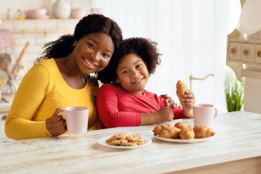 Afrikalı anne ve küçük kız mutfakta bir şeyler atıştırıyor, kruvasan yiyor ve birlikte kakao içiyor, siyahi aile evde vakit geçiriyor, elinde fincanlarla kameraya bakıyor, bedava uzay boşluğu seyrediyor.