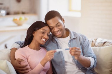 Evde bebeklerini bekleyen pozitif hamilelik testiyle gülümseyen anne ve baba. Genç siyahi adam mutlu hamile karısına evde sarılıyor, mutlu haberleri birlikte öğreniyor.