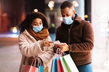 Perakende satış, Teknoloji, Koruma ve Salgın Konsepti. Tıbbi maske takan, cep telefonu kullanan, ekranı gösteren, alışveriş torbası tutan, akşam vakti yürüyen siyah bir çiftin portresi.