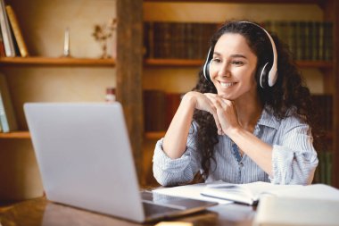 Sanal konferans. Kablosuz kulaklıklı neşeli kız öğrencinin portresi masada oturuyor, öğretmeniyle çevrimiçi ders alıyor ve dizüstü bilgisayarına bakıyor. Ofiste çalışan gülümseyen kadın.