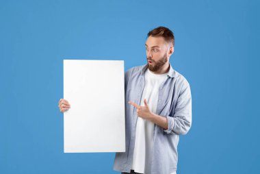 Mavi stüdyo arka planındaki beyaz boş posteri gösteren yeni nesil bir adam. Reklam tasarımın için bir model. Yakışıklı beyaz adam, boş afişi kopyalama alanıyla gösteriyor