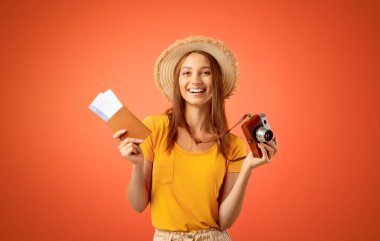 Yazlık şapkalı, elinde kamera ve pasaportu olan gülümseyen genç bir kadın.