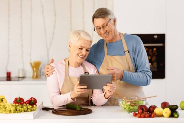 Neşeli Yaşlı Çift Dijital Tablet Yemek Pişiriyor, İnternette Yemek Tarifleri Arıyor Modern Mutfakta Yemek Hazırlıyor. Aşçılık Uygulaması Konsepti.