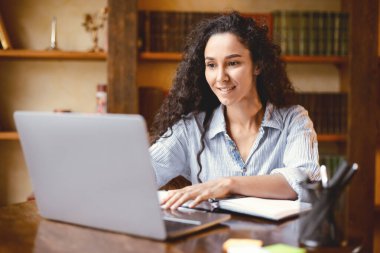 Proje yapıyor. Dizüstü bilgisayar kullanan, klavyeye yazı yazan, masada oturan kıvırcık bir genç kadının portresi. Uzaktan çalışan ya da evde internetten çalışan bir bayan yönetici.
