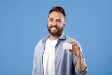 Mutlu genç adam mavi stüdyo arka planında tasarım için yeri olan boş bir kartvizit gösteriyor. Gülümseyen sakallı adam reklam için elinde boş reklam kartı tutuyor.