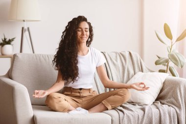 Rahatlama, stres atma konsepti. Uzun kıvırcık saçlı barışçıl esmer kadın oturma odasındaki kanepede oturmuş meditasyon yapıyor, fotokopi çekiyor. Olumlu genç bayan evde hafta sonunun tadını çıkarıyor.