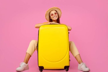 Yaz şapkalı ve güneş gözlüklü güzel kız pembe stüdyo arka planında bavuluyla poz veriyor.