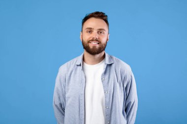 Mavi stüdyo arka planında poz veren ve gülümseyen rahat giyinen neşeli genç adamın portresi. Tişörtlü, mutlu yüz ifadesi olan olumlu sakallı bin yıllık bir adam.