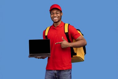 Sipariş ver. Elinde dizüstü bilgisayarla gülümseyen siyah bir teslimatçının portresi. Kırmızı üniformalı ve sarı termo çantalı boş ekranı işaret ediyor. Mavi stüdyo arka planında duruyor.