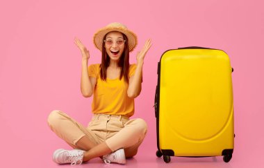 Şapkalı ve güneş gözlüklü duygusal kız valizinin yanında oturuyor, seyahati için heyecanlı, pembe arka plan.
