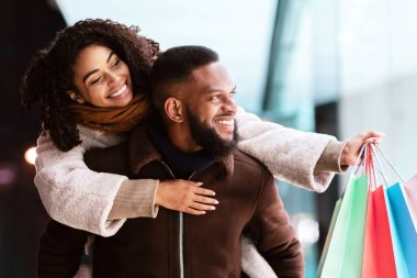 Sağlıklı bir ilişki. Gülümseyen Afrikalı Amerikalı adam, elinde alışveriş torbalarıyla gezen mutlu karısına sırtına biniyor. Bir çift dükkanın camına bakıyor, akşam vakti merkezde yürüyor.