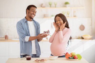 Hamilelik sırasında tatlılar. Genç siyah adam mutfaktaki hamile sevgilisine lezzetli bir pasta veriyor. Yakışıklı Afrikalı Amerikalı koca sevgili karısına tatlı ikram ediyor.