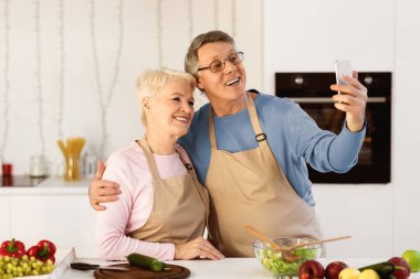 Neşeli son sınıf çifti cep telefonuyla selfie çekiyor. Evde modern mutfakta birlikte yemek pişiriyorlar. Yaşlı Eşler Akıllı Telefon Konsepti Kullanarak Yemek Hazırlığı Yapıyor