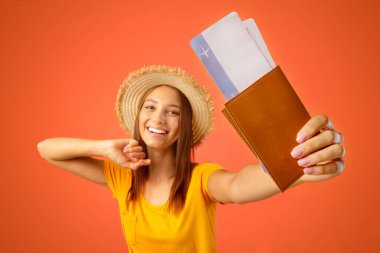 Pasaportu ve uçuş biletleri olan, sarı stüdyo geçmişi olan, yaz şapkalı neşeli genç bir kadın. Pasaportunu uçak biletleriyle gösteren mutlu bayan gezgin, neşesini arkadaşlarıyla ve takipçileriyle paylaşıyor.