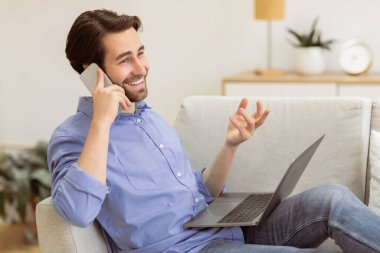 Cep telefonuyla konuşan neşeli bir adam dizüstü bilgisayarla evde oturuyor. Keyifli Telefon Konuşması Yapan Adam. Modern İletişim ve Serbest Yaşam Tarzı. Yan Görünüm