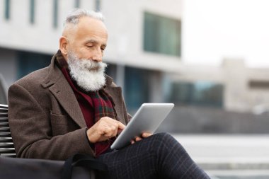 Beyaz saçlı şık bir adam ofis binasının yanındaki bankta oturuyor, dijital tablet kullanıyor, ajandasını kontrol ediyor, kopyalama alanını kopyalıyor. Yaşlı sakallı adam sokakta ped kullanıyor, dinlenirken web sörfü yapıyor.