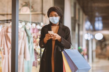 Alışveriş için modern cihazlar ve mağazadan satış ve Kara Cuma ile ilgili mesajlar. Koruyucu maskeli genç Afrikalı Amerikalı kadın alışveriş çantalarını tutuyor ve sosyal medyada yazmak için telefon kullanıyor.