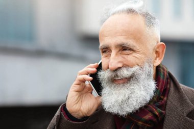 Sıcak kıyafetler içinde, sokakta yürürken cep telefonuyla konuşan neşeli, kıdemli bir adamın yakın plan portresi. Yakışıklı, sakallı, yaşlı iş adamı dışarıda akıllı telefondan konuşuyor.