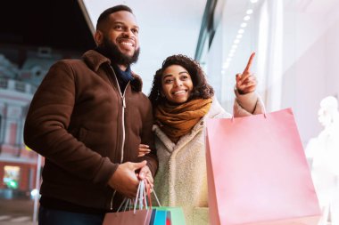 Satış ve İndirim. Neşeli kıvırcık siyah kadın alışveriş merkezinin camını işaret ediyor, vitrini gösteriyor, akşamları alışveriş merkezinin yakınında erkeğiyle yürüyor, elinde renkli torbalarla alışverişler yapıyor.