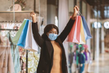 Covid-19 salgını ve uzun zamandır beklenen alım sırasında Kara Cuma 'da alışveriş ve sezonluk satış. Renkli alışveriş torbalarıyla koruyucu maskeli neşeli Afro-Amerikalı kadın kazanma duygularını ifade ediyor.