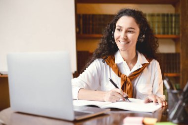 Ödev yapıyorum. Gülümseyen kıvırcık kadının portresi elinde kalemle oturuyor, not alıyor, dizüstü bilgisayarını kullanıyor ve ona bakıyor. Yetişkin kadın karantina sırasında online kurs izliyor.
