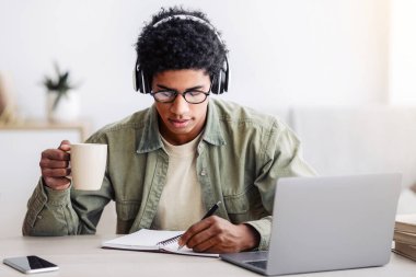 İnternetteki bilgisayar dersinde kahve içen meşgul bir genç. Not defterine, eve bilgi yazıyor. Siyah erkek öğrenci evde eğitim görüyor, sanal sınıfta çalışıyor, öğretmenini dinliyor.