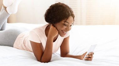 Rahat Afro-Amerikan genç bayan akıllı telefonuyla yatağa uzanıyor, sosyal medya ya da blog aracılığıyla arkadaşlarıyla sohbet ediyor, güzel uygulamalar kullanıyor, hafta sonları evde dinleniyor, uzay kopyaları kopyalıyor.