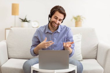 Kulaklıklı neşeli adam dizüstü bilgisayarla video görüşmesi yapıyor. Uzaktan çalışan meslektaşıyla konuşuyor. Evdeki koltukta oturuyor. Video Konferansı, Modern İletişim, Çevrimiçi İş Eğitimi Konsepti