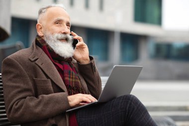 Modern dizüstü bilgisayarı olan yaşlı sakallı bir iş adamı bankta oturuyor, telefonla konuşuyor, boş bir alanda. Tarz sahibi, yaşlı, gri saçlı bir adam iş merkezinin yanında sokakta oturuyor ve internette çalışıyor.