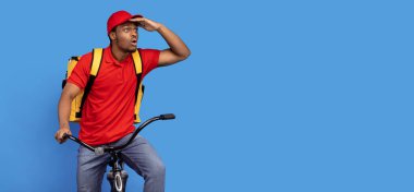 Hızlı Teslimat Servisi. Duygusal olarak şaşırmış kırmızı üniformalı bir kurye bisiklet sürüyor, termo çanta takıyor ve mavi arka planda, pankartta, pankartta izole edilmiş fotokopi alanına bakıyor.