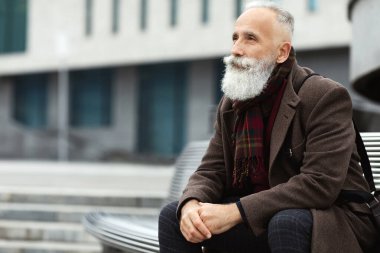Gri saçlı ve uzun sakallı yakışıklı bir adam ofisin yanındaki bankta oturuyor, kalın bir ceket giyiyor, deri çanta giyiyor, öğle arasında dinleniyor, fotokopi odasına bakıyor.