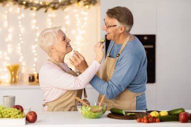 Happy Senior Couple Feeding Each Other Cooking Together In Modern Kitchen At Home. Christmas Dinner Preparation, New Year Celebration And Holidays Recipes Concept