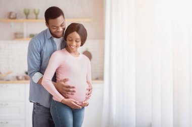 Beklenen Afro-Amerikan kadın ve mutlu erkek arkadaşı evde pencerenin önünde kucaklaşıyorlar. Çekici siyahi adam hamile eşinin karnına dokunuyor, romantik anlar yaşıyor.