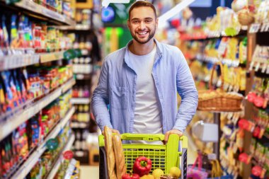 Market alışverişi. Gülümseyen mutlu sakallı adam Hypermarket, kapalı alanda alışveriş arabasıyla dikiliyor. Neşeli adam dükkandan sağlıklı yiyecekler alıyor, kameraya bakıyor ve poz veriyor.