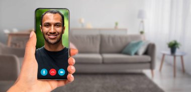 Yakışıklı bir adamla görüntülü konuşma yapan bir akıllı telefon tutan kişi. Arka planda kanepesi ve doğal ışığı olan rahat bir oturma odası var. İletişim için rahat bir atmosfer yaratıyor..