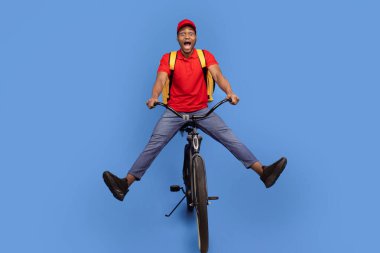 Hızlı Teslimat Servisi. Kırmızı şapkalı ve üniformalı komik Afro-Amerikan kurye bisiklete biniyor ve mavi arka planda ısıl çanta giyiyor. Tecrit sırasında sevkiyat servisi