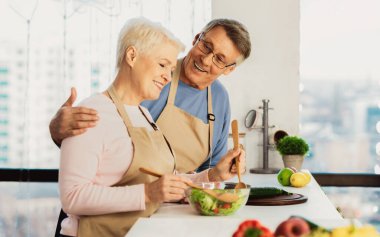 Son sınıf mutlu eşlerin modern mutfaklarda birlikte yemek pişirmelerinin yan görüntüsü. Yaşlı koca ve eş evde ayakta durarak akşam yemeği için sağlıklı salata yapıyorlar. Beslenme ve aile tarifleri