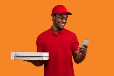 Fast Service ve en iyi yemek teslimatı. Kırmızı üniformalı ve şapkalı gülümseyen siyah kurye restorandan pizza kutularını tutuyor ve turuncu stüdyo arka planında izole edilmiş akıllı telefon kullanıyor.