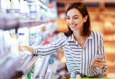 Alışverişe. Gülümseyen Genç Kadın markette ayakta durup telefon kullanıyor ve yiyecek alışverişi yapıyor. Akıllı telefonlu bir bayan müşteri mandıra bölümündeki dondurucudan süt alıyor.
