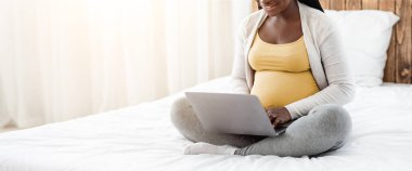 Evde dizüstü bilgisayar kullanan hamile bir kadın, uzaktan çalışıyor, internete bakıyor ya da internetten alışveriş yapıyor, bilgisayarla rahat bir yatakta oturan, klavyede yazan, boşluğu kopyalayan Afrikalı bir kadın bekliyor.