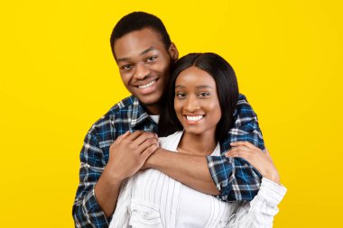 Aşk, randevu, ilişki, aile ve birlikte mutlu olmak. Gülümseyen bin yıllık Afro-Amerikan erkeği eşine sarılıyor ve kameraya bakıyor, sarı arka planda izole edilmiş, stüdyo çekimi, serbest alan.